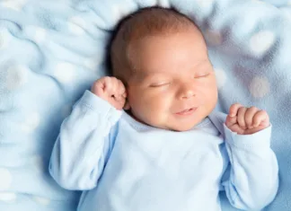 20 najpogostejših novodobnih imen za dečke v Sloveniji Newborn Baby Sleeping Smiling. Cute Infant Child in Wrap Bodysuit. New Born Little Boy smile in Blue Clothes lying on Soft Blanket. Small Kid Face Close up Portrait holding Hands First