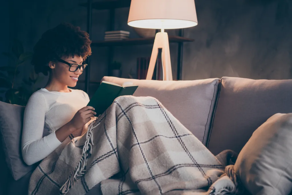 Profile side photo of positive dreamy afro american girl sit divan read paperbook, enjoy fiction story covered by plaid checkered blanket in room apartment