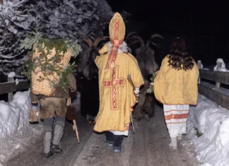 Miklavževanja 2024: Kam v Sloveniji na Miklavžev obisk? This image depicts a group of Krampus figures walking through a snowy village in Austria. The figures are dressed in traditional Krampus costumes, with horns, fur, and bells.