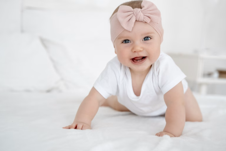 A baby girl is smiling at home on a white bed with a bow on her head, a happy healthy child of six months is laughing.