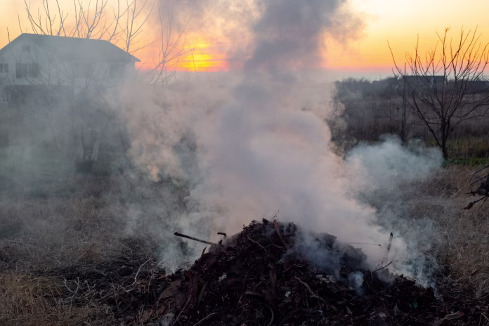 A pile of rubbish from leaves and branches burns in a rural field, smoke rises into the evening sunset sky. Cleaning the garden.