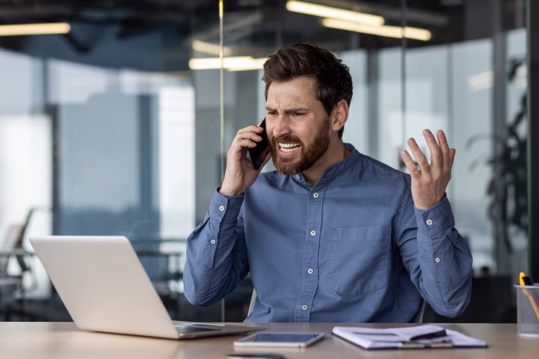 Angry and irritated young businessman sitting in the office at the desk and talking on the phone while waving his hands.