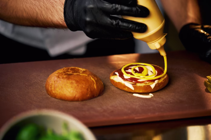 Close up view of hamburger, preparing with ketchup and mayo sauce on cutting board. Crop view of chef's hands in gloves, pouring mustard on grilled buns, making cheeseburger. Concept of fast food.