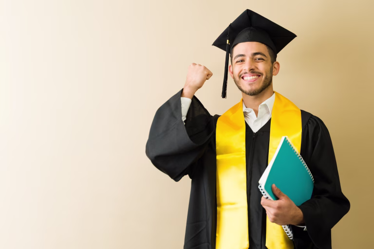Excited male graduate raising fist in celebration, holding notebook, wearing graduation gown and smiling, ready for future career