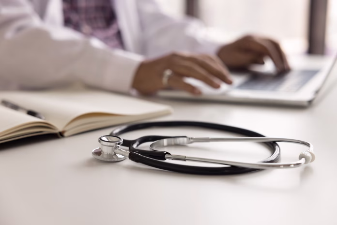 Medical practitioner stethoscope on white table surface close up shot with hands of male doctor typing on laptop computer in blurred background. Physician workplace cropped shot