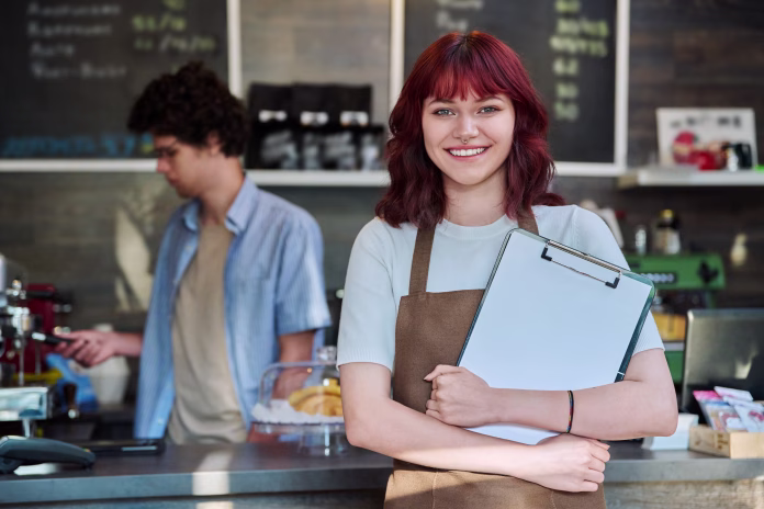 Portrait of young smiling female waitress in an apron holding clipboard in hands, standing in front of counter in coffee shop. Food service occupation, part time job for students, staff, work concept