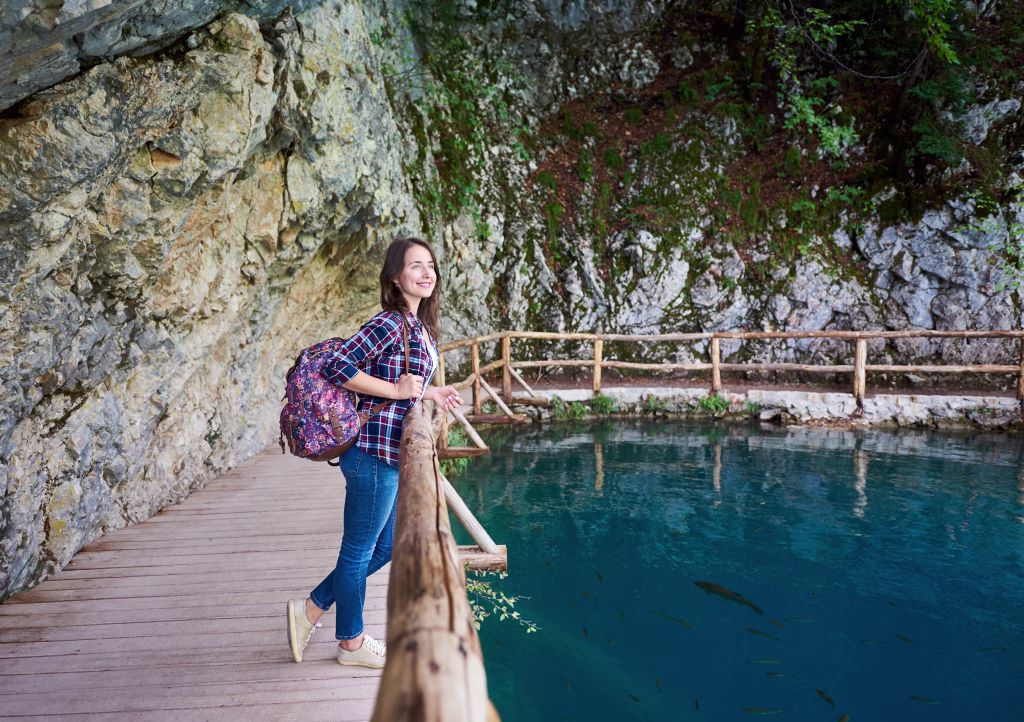 Slim pretty smiling tourist girl with backpack standing on wooden pathway under green tree enjoying beautiful view of clean blue lake lit by summer sun. Plitvice National Park, Croatia, Europe.