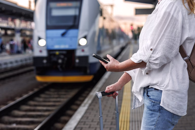 Woman with suitcase is using smartphone and mobile app to purchase online transport ticket. Tourist waiting for train at the station. Solo travel