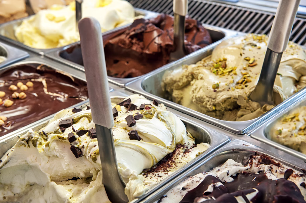 Close up of trays of different ice cream in assorted flavors displayed in a store window in metal trays with scoops for takeaway summer snacks