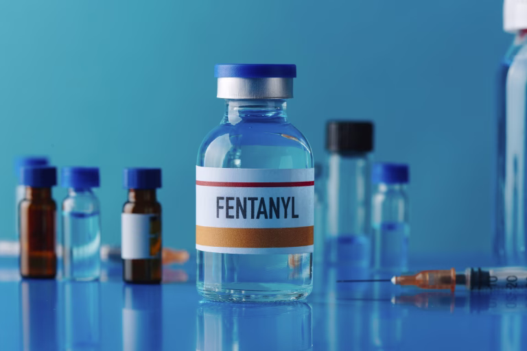 closeup of a simulated vial of fentanyl on a blue table next to a syringe and some other different vials
