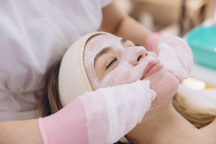 Gloved beautician spreading white facial mask across client's skin during professional skincare treatment in spa setting