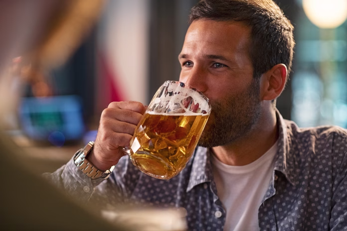 Handsome young man drinking a pint of beer while looking away. Cheerful mid man in casual clothing feeling relaxed while enjoying draft beer in bar. Middle aged guy take a sip from his drink at pub during the night.