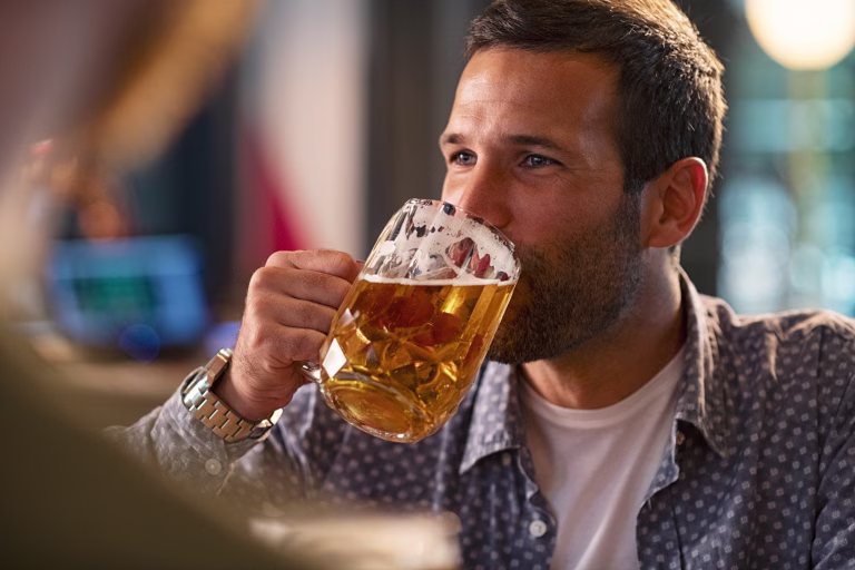 Handsome young man drinking a pint of beer while looking away. Cheerful mid man in casual clothing feeling relaxed while enjoying draft beer in bar. Middle aged guy take a sip from his drink at pub during the night.