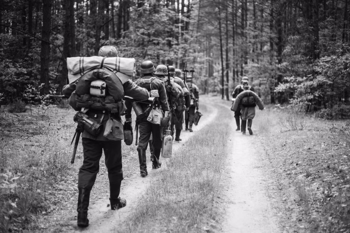 Re-enactors Dressed As German Infantry Soldiers In World War II Marching Walking Along Forest Road In Summer Day. Photo In Black And White Colors.