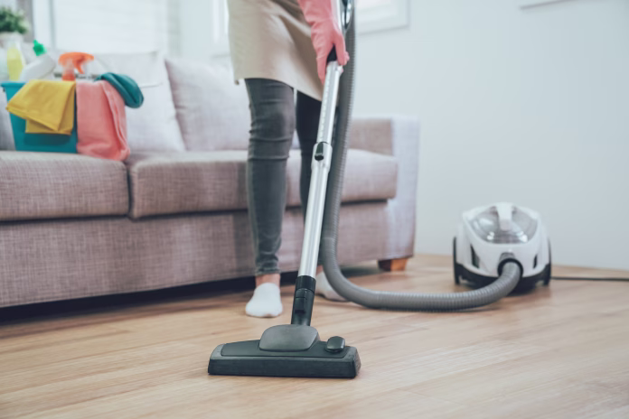 asian woman using vacuum cleaner in the living room. housekeeper wearing pink protective rubber gloves cleaning the wooden floor in the house. full of clean supplies in bucket on comfortable sofa
