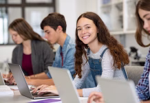 Tukaj smo – za prave finančne zvezde! Happy young woman working on laptop and looking at camera in classroom. Portrait of smiling university student in library use computer for a research. Satisfied college student looking at camera while sitting in a row with classmates studying together.