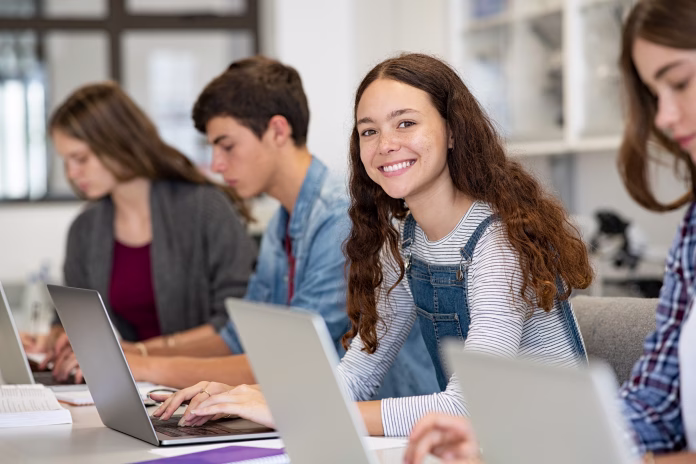 Happy young woman working on laptop and looking at camera in classroom. Portrait of smiling university student in library use computer for a research. Satisfied college student looking at camera while sitting in a row with classmates studying together.