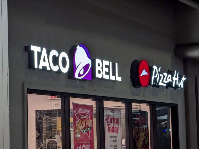 Honolulu - December 6, 2022: Taco Bell and Pizza Hut storefront in a strip mall at night. The storefront has a black awning with white lettering and logos. The windows display advertisements and a hiring sign. The parking lot and other stores are visible in the background.