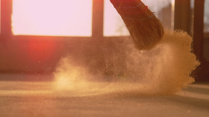 LENS FLARE, CLOSE UP, LOW ANGLE: Dust gets swept up into air as an unrecognizable person cleans the construction site floor. Contractor sweeps the ground of a construction site with a straw broom