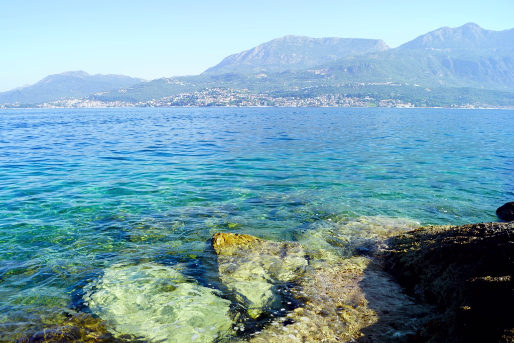 Vibrant seascape from the coast of Montenegro: clear blue waters of the sea and a coastal town at the foot of the mountains in the distance.