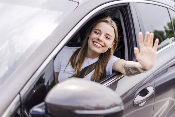 Young woman happily waves from her car window on a sunny road trip, embodying the spirit of summer and adventure