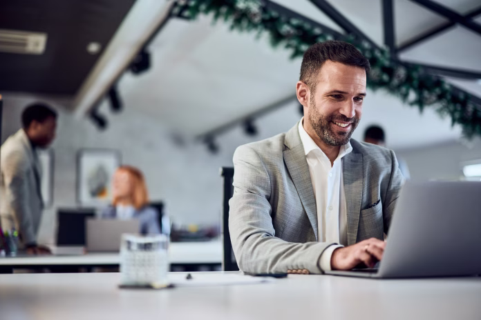 A happy businessman working surrounded his colleagues at the office, typing an email over the laptop.