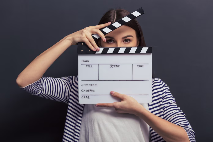 beautiful-young-girl-in-casual-clothes-is-covering-her-face-with-a-clapperboard-and-looking-at-camera-standing-against-blackboard-stockpack-istock Beautiful young girl in casual clothes is covering her face with a clapperboard and looking at camera, standing against blackboard