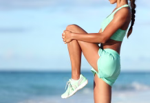 Kako dolgoročno zaščititi svoja kolena? Fitness runner body closeup doing warm-up routine on beach before running, stretching leg muscles with standing single knee to chest stretch. Female athlete preparing legs for cardio workout.