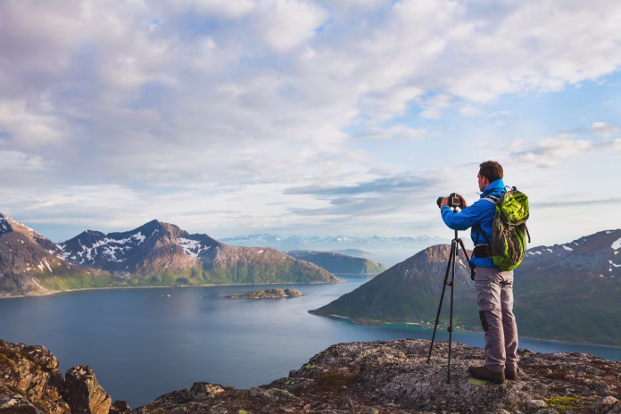 landscape photographer working with tripod and dslr camera in beautiful wild nature, standing with backpack on top of mountain