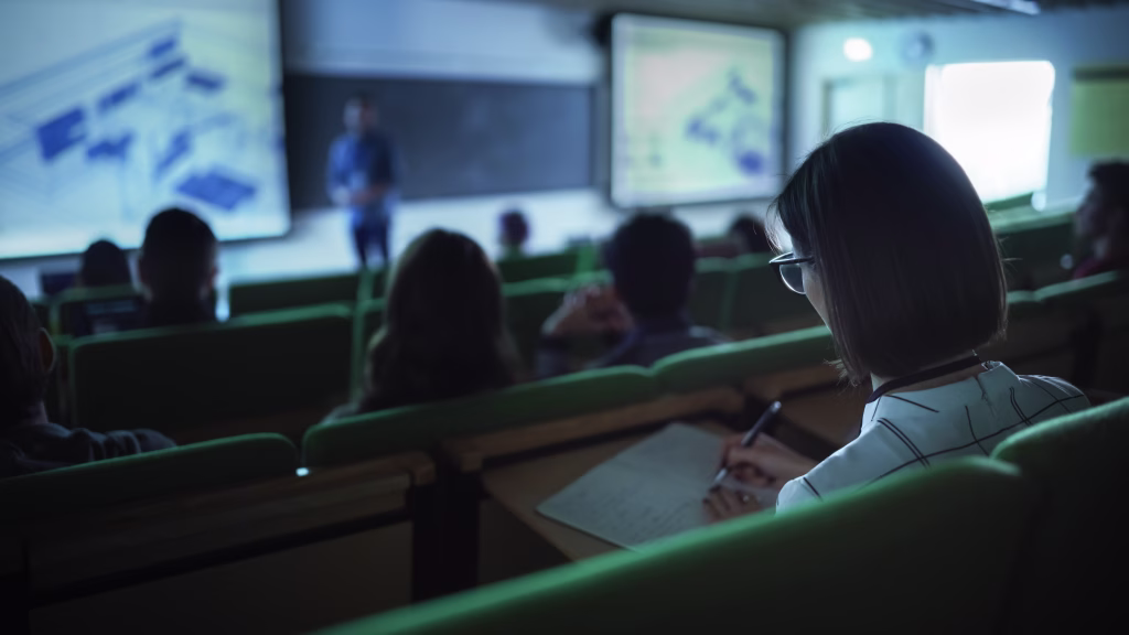 Over the Shoulder Footage of a Female Student Taking Down Notes in a Notebook, Sitting in College Auditorium on a Lecture. Young Woman with Short Hair Wearing Glasses and Studying in University