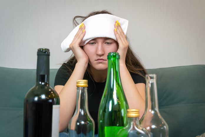 Young woman with a white towel on her forehead from a headache sits in front of empty bottles of alcohol. Concept of Monday morning, morning after drinking alcohol