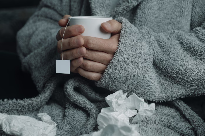 an ill man at home, wearing a fluffy gray house robe, surrounded by used tissues, warms up with a cup of hot herbal tea