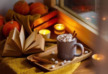 Knjige, ki jih moraš prebrati to jesen! Mug of cocoa or hot chocolate with marshmallows next to the window with candles, pumpkins, book and warm blanket. Cozy home atmosphere in rainy autumn day. Нygge lifestyle concept