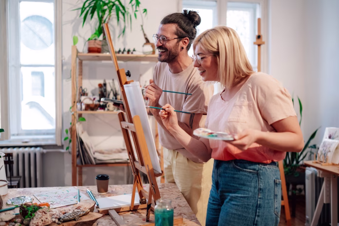 Smiling young creative painters standing at art studio and painting together on easel. Affectionate happy artist couple crating unique artwork on canvas at creative atelier and bonding. Couple goals.