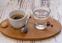 Ali veste, zakaj vam ob kavi postrežejo kozarec vode? Ni namenjena pitju, kot mnogi mislijo … White cup of Espresso and a glass of water disposed on a wooden plate