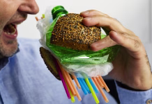 Kako zmanjšati svojo izpostavljenost mikroplastiki? – Upoštevaj TE nasvete za zdravje A man eats a burger made from a tasty bun with sesame and poppyseed filled with different types of disposable plastic. The concept of disposable plastic as a danger for ecology and human health.