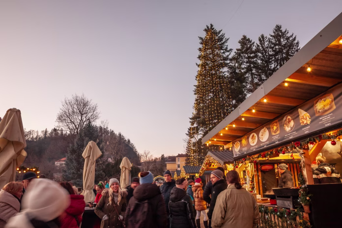 Bled, Slovenia - December 17, 2024: Early evening at the lakeside town of Bled finds locals and tourists mingling around the annual Christmas market. Timber kiosks, trimmed with garlands and fairy lights, offer traditional Slovenian street food, hot mulled wine, pastries and cheeses. A majestic fir tree wrapped in hundreds of tiny bulbs rises above the crowd, its reflection glimmering off nearby umbrellas closed for the day. Bundled visitors chat, laugh and warm their hands on steaming cups, embodying the Alpine concept of cosy winter socialising. The candid scene speaks to European Advent traditions, sustainable small town tourism and the vibrant holiday spirit that transforms Bled each December.