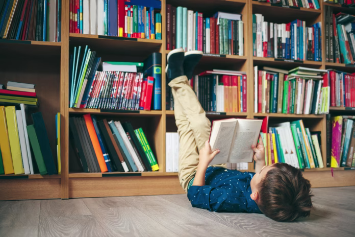 boy-laying-on-the-floor-with-the-feet-up-reading-a-book-against-multi-colored-bookshelf-in-library-education-knowledge-bookstore-lecture-pupil-holds-a-book-in-his-hands-stockpack-istock Boy laying on the floor with the feet up, reading a book against multi colored bookshelf in library. Education, Knowledge, Bookstore, Lecture. Pupil holds a book in his hands.