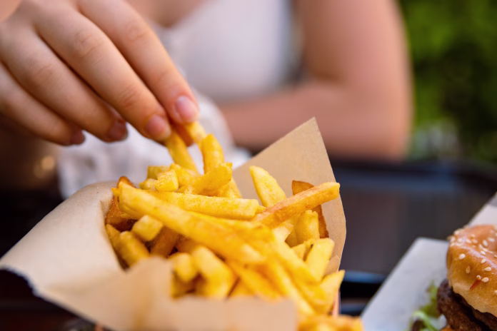 Closeup of young womans hand grabbing a crispy golden fries. Enjoying potato chips with a juicy burger in fast food restaurant on summer day. Selective focus