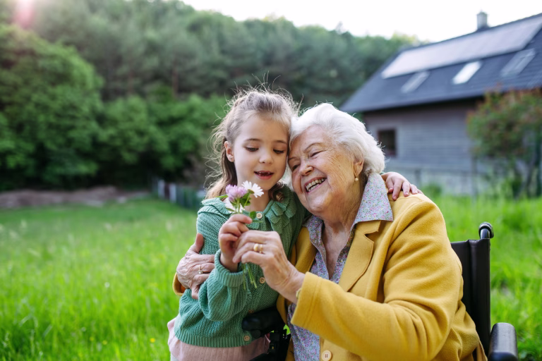 Granddaughter spending time with elderly grandma, picking widlflowers. Senior lady in wheelchair spending time in nature. Weekend in cottage.