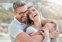 Zakaj ženske živijo dlje? Happy and loving mature caucasian couple enjoying a romantic date at the beach together on a sunny day. Cheerful affectionate husband hugging arm around wife while bonding on vacation outdoors