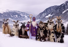 Kdo je bil pravi Miklavž? In a snowy mountain setting, a group of people dressed in traditional costumes stands together, including St. Nicholas and mystical companions, celebrating a vibrant winter festival.