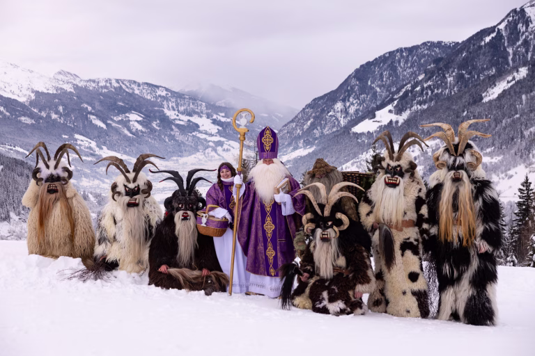 In a snowy mountain setting, a group of people dressed in traditional costumes stands together, including St. Nicholas and mystical companions, celebrating a vibrant winter festival.