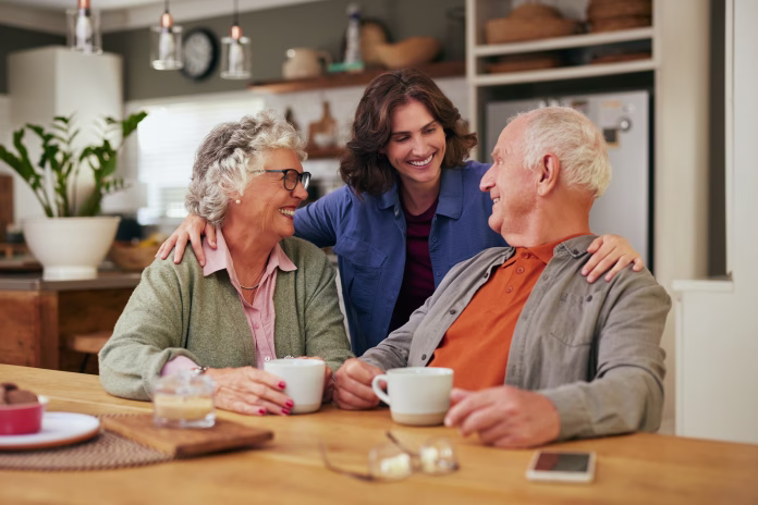 Senior father and old mother sharing tea and heartfelt conversation with daughter at home at tea time. Elderly parents and beautiful daughter bonding over tea and discussion. Warm daughter embracing from behind her joyful parents while sitting at table and having a good conversation.