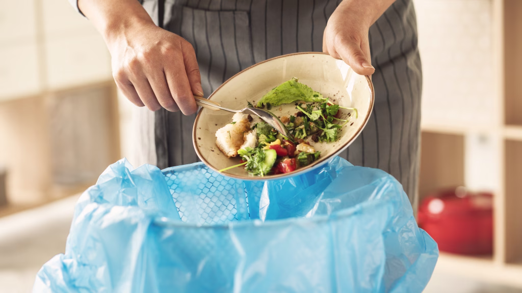 Woman scraping leftover food from plate into trash bin in kitchen