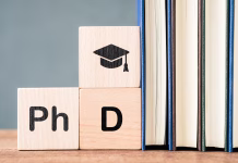 Doktorat: DA ali NE? Wooden blocks displaying PhD text and graduation cap symbol placed beside stacked books, representing advanced academic study, higher education goals, research degree, and scholarly achievement
