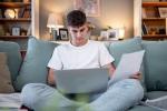 Young man sitting comfortably on a sofa in his living room, working on his laptop while reviewing documents, demonstrating focused concentration in a home office setting