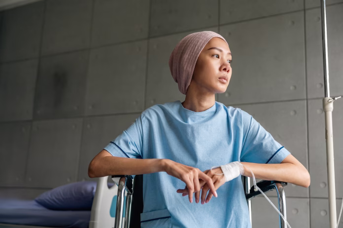 A cancer patient sits in a wheelchair with an IV drip looking stressed and sad. Asian Woman receiving IV therapy in a wheelchair showing visible signs of stress and depression