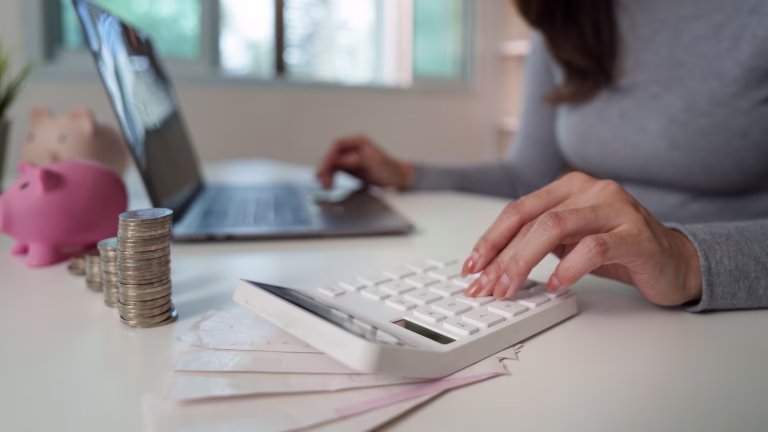 A woman calculates her expenses using a calculator while managing her finances on a laptop, emphasizing budgeting skills.