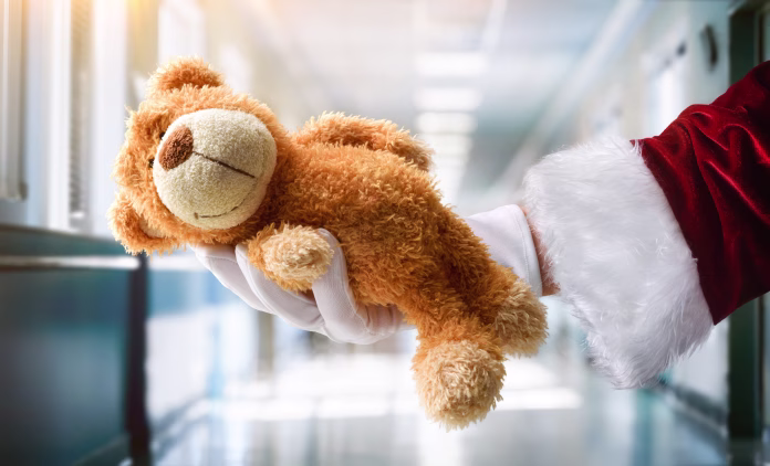 Concept of giving toys to hospitalized children at Christmas with Santa Claus's hand wearing a white glove holding a teddy bear in a hospital corridor.